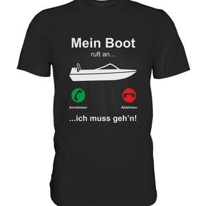 Könnte beinhalten: Schwarzes T-Shirt mit weißem Text, der "Mein Boot ruft an..." lautet und einem Cartoon-Bild eines Bootes mit einem Telefonsymbol darüber. Der Text unter dem Bild lautet "Annehmen" und "Ablehnen" mit grünen bzw. roten Telefonsymbolen. Der Text unter den Symbolen lautet "...ich muss geh'n!"