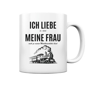 May include: White ceramic mug with a black design that says "Ich liebe es, wenn meine Frau mich zu meiner Modelleisenbahn lässt."  The design also includes a black and white illustration of a steam train.