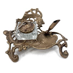 May include: Antique brass inkwell set with a clear glass ink pot and a pen holder. The inkwell has an ornate, scrolled design and a small chain attached to the lid. The pen is a bronze color.