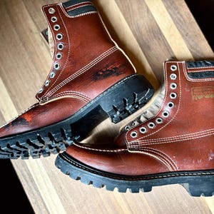 Könnte beinhalten: Ein Paar braune Leder-Arbeitsstiefel mit schwarzen Sohlen. Die Stiefel haben mehrere Ösen für Schnürsenkel und weiße Nähte. Die Sohlen haben ein tiefes Profil. Die Stiefel stehen auf einer Holzoberfläche.
