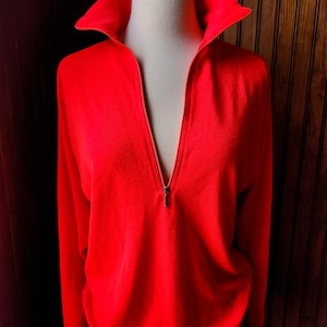 May include: A bright red long-sleeved sweater with a zip up the front. The sweater has a high collar and is made of a soft, knitted fabric.