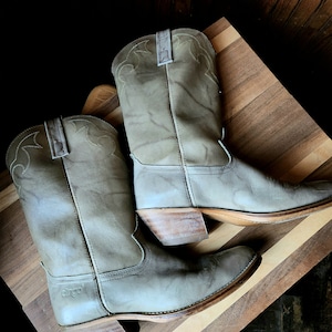 May include: A pair of gray leather cowboy boots with a pointed toe and a low heel. The boots have a stitched design on the top and the word "Dingo" is embossed on the side of one boot.