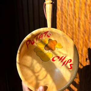 Puede incluir: Una bandeja de madera vintage con asa, con las palabras "POTATO CHIPS" en letras rojas. La bandeja tiene una ilustración de dibujos animados de una patata frita con sombrero. El asa está sujeta con una cuerda rosa.