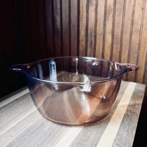 Puede incluir: Un cuenco de vidrio transparente de color ámbar con dos asas. El cuenco está sobre una tabla de cortar de madera con rayas claras y oscuras. El fondo es una pared de madera.