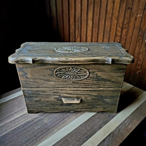 Peut inclure: Une boîte en bois avec un motif floral sculpté sur le couvercle et un tiroir. La boîte est posée sur une surface en bois rayée.