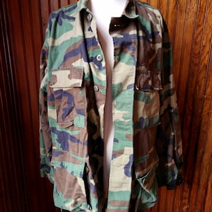 Könnte beinhalten: Eine Jacke im Tarnmuster mit Knopfleiste und mehreren Taschen. Die Jacke ist offen und hängt an einem Mannequin.