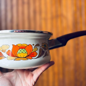 May include: Cream-colored saucepan with a floral design in orange, yellow, and brown. The pot has a dark brown handle and a silver rim. A vintage kitchenware item.