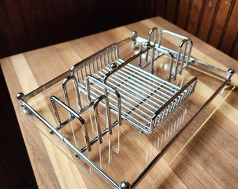 Op de afbeelding: Een verchroomd metalen afdruiprek met een transparante acrylbasis. Het rek heeft meerdere compartimenten voor het opbergen van serviesgoed, bestek en andere items.
