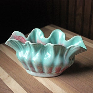 May include: A turquoise and pink ceramic planter with a ruffled edge. The planter is shaped like a shell and has a glossy finish.