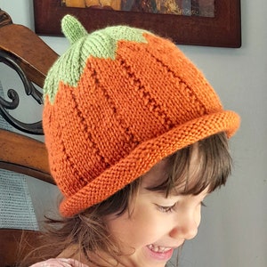 May include: A child wearing a knitted orange and green pumpkin hat. The hat has a pointed top and a ribbed texture. The child is smiling and looking down at a book.