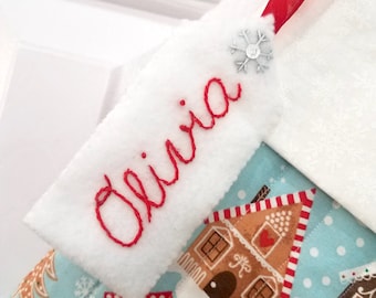 May include: White stocking with a red ribbon and a snowflake embellishment. The stocking has a white felt tag with the name "Olivia" embroidered in red thread.