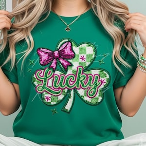 Op de afbeelding: Groen T-shirt met een klaverontwerp, een roze strik en het woord "Lucky". De klaver heeft een groen-wit geruit patroon. Het T-shirt heeft een effen groene kleur. Perfect voor St. Patrick's Day.