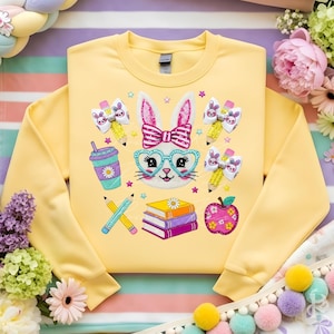 Könnte beinhalten: Ein gelbes Sweatshirt mit einem Cartoon-Hasen-Design. Das Design umfasst Brille, Schleife, Bleistifte, Bücher, ein Getränk und Schleifen. Das Sweatshirt ist mit farbenfrohen Stickereien verziert, ideal für Ostern oder den Frühling.