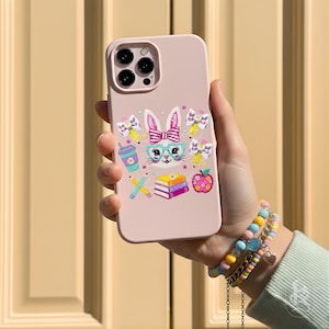 Puede incluir: Una funda de tel&eacute;fono rosa claro con un conejito de dibujos animados con gafas y un lazo. La funda tiene ilustraciones coloridas de &uacute;tiles escolares, incluyendo l&aacute;pices, libros y una manzana. La funda del tel&eacute;fono se sostiene en una mano, con una pulsera colorida en la mu&ntilde;eca.