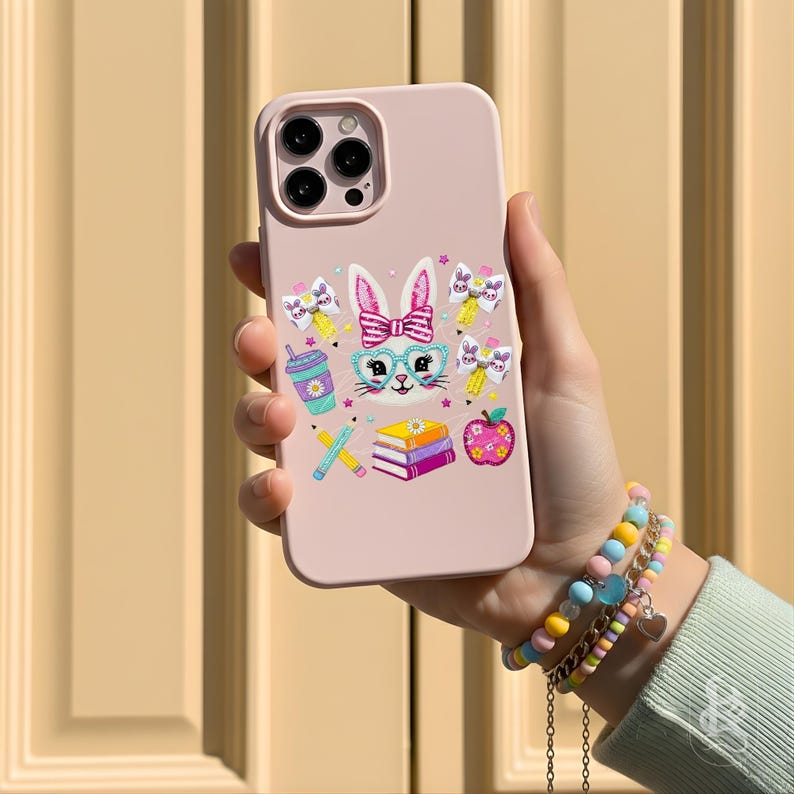 Puede incluir: Una funda de tel&eacute;fono rosa claro con un conejito de dibujos animados con gafas y un lazo. La funda tiene ilustraciones coloridas de &uacute;tiles escolares, incluyendo l&aacute;pices, libros y una manzana. La funda del tel&eacute;fono se sostiene en una mano, con una pulsera colorida en la mu&ntilde;eca.
