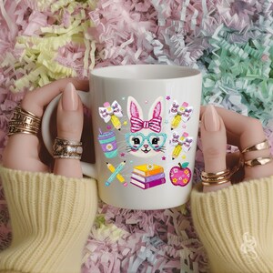 Puede incluir: Taza de cer&aacute;mica blanca con un dise&ntilde;o de conejo de dibujos animados, que incluye gafas, una pajarita y gr&aacute;ficos escolares. El dise&ntilde;o presenta l&aacute;pices, libros, una manzana y una bebida. La taza la sostiene una persona que lleva un su&eacute;ter amarillo y varios anillos.