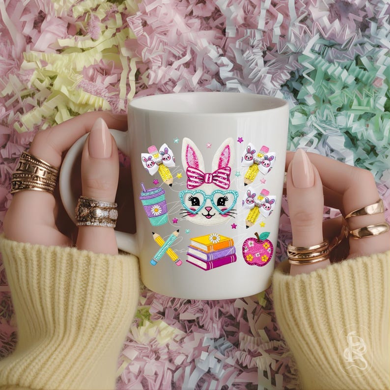 Puede incluir: Taza de cer&aacute;mica blanca con un dise&ntilde;o de conejo de dibujos animados, que incluye gafas, una pajarita y gr&aacute;ficos escolares. El dise&ntilde;o presenta l&aacute;pices, libros, una manzana y una bebida. La taza la sostiene una persona que lleva un su&eacute;ter amarillo y varios anillos.