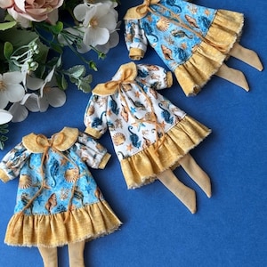 May include: Three doll dresses with a seashell and seahorse print on a blue background. Each dress has a yellow ruffled skirt and a matching bow at the collar. The dresses are arranged with a floral arrangement in the background.