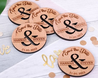 May include: Five wooden save the date circles with the text "Save the Date" and an ampersand symbol. The names "Hannah" and "Steven" are on each circle, along with the date "21.05.2022".