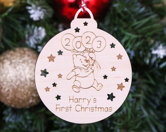 Op de afbeelding: Een houten kerstversiering in de vorm van een bal met een Winnie de Poeh-figuur die ballonnen vasthoudt met de tekst 2023. De versiering is versierd met sterren en de tekst "Harry's First Christmas".