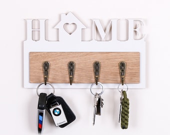 Puede incluir: Porta llaves de madera blanco con la palabra "HOME" en letras recortadas. El porta llaves tiene cuatro ganchos y un corazón recortado en la forma de la casa. Hay llaves colgando de los ganchos.