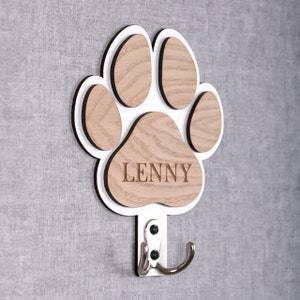 May include: White wooden paw print coat hook with the name "Lenny" engraved on the bottom. The hook is attached to the paw print and is silver.
