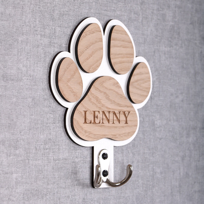 May include: White wooden paw print coat hook with the name "Lenny" engraved on the bottom. The hook is attached to the paw print and is silver.