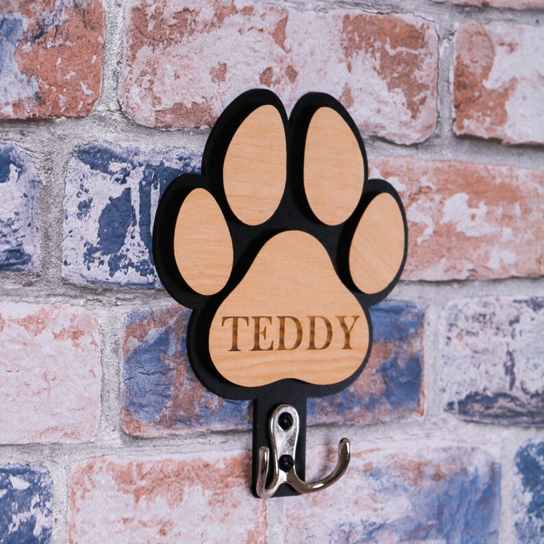 May include: Wooden paw print shaped hook with a black outline and the name "TEDDY" engraved on it. The hook is attached to a brick wall.