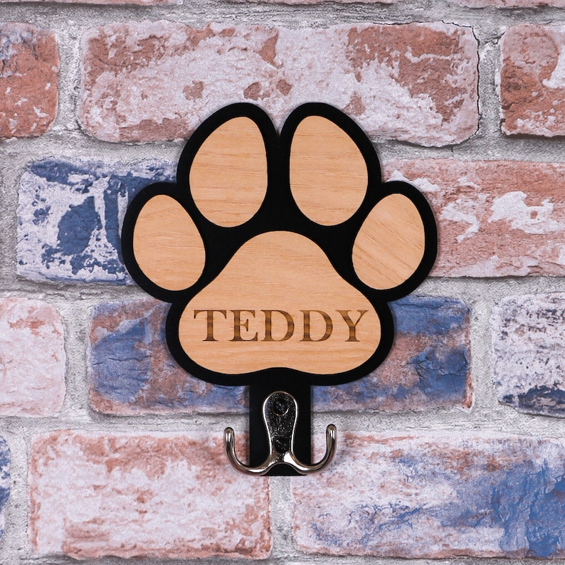 May include: Black paw print shaped wooden hook with the name "Teddy" engraved on it. The hook is mounted on a brick wall.