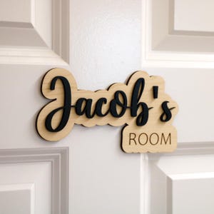 May include: A wooden door sign with the text "Jacob's Room" in black lettering. The sign is made of light wood and has a slightly curved shape.