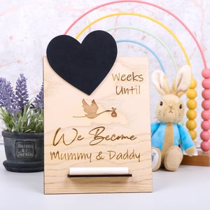 Puede incluir: Un tablero de cuenta atrás de madera con una pizarra en forma de corazón negro, una cigüeña que lleva un bebé y el texto "Semanas Hasta Que Nos Convertimos en Mamá & Papá".