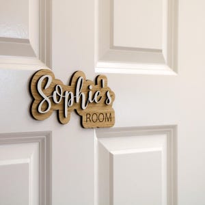 May include: A wooden door sign with the text "Sophie's Room" in white lettering. The sign is made of light brown wood and has a slightly distressed look.