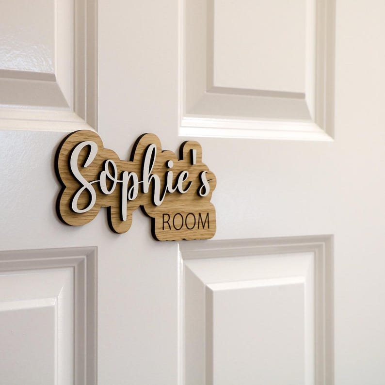 May include: A wooden door sign with the text "Sophie's Room" in white lettering. The sign is made of light brown wood and has a slightly distressed look.