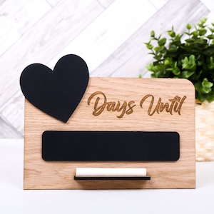 Könnte beinhalten: Ein hölzerner Countdown-Schild mit einem schwarzen Schieferherz und einem kleineren Schieferrechteck. Das Schild sagt "Days Until" und hat einen kleinen Kreidehalter.