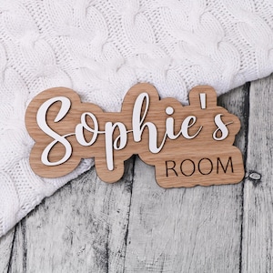 May include: A wooden door sign with the text "Sophie's Room" in white lettering. The sign is made of light brown wood and has a slightly rounded shape.