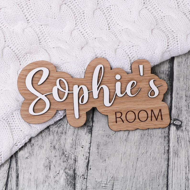 May include: A wooden door sign with the text "Sophie's Room" in white lettering. The sign is made of light brown wood and has a slightly rounded shape.