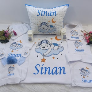 Könnte beinhalten: Ein Baby-Geschenkset in Weiß und Blau. Das Set enthält ein Kissen, ein Lätzchen, einen Strampler, eine Mütze und Fäustlinge. Die Artikel sind mit einem niedlichen Cartoon-Bären verziert, der auf einer Wolke schläft, und der Name "Sinan" ist auf jedem Artikel gedruckt.