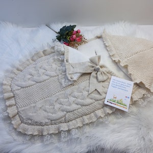 Può includere: Un sacco a pelo per neonati in maglia color crema con un bordo in pizzo e un fiocco. Il sacco è piegato e ha un piccolo biglietto con il testo "Wir wünschen, dass Sie es in gesunden und glücklichen Tagen verwenden." Il biglietto ha anche il testo "Baby und Kleinkind" e un logo.
