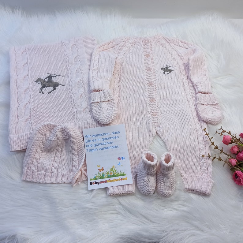 May include: A pink knitted baby outfit set with a hat, booties, and a one-piece with attached mittens. The outfit features a cable knit pattern and a small embroidered horse design. The set comes with a card that says "Wir w&uuml;nschen, dass Sie es in gesunden und gl&uuml;cklichen Tagen verwenden."