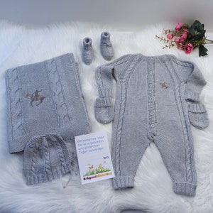 May include: A grey knitted baby outfit set with a hat, booties, and a sleepsuit. The sleepsuit has a cable knit pattern and a small embroidered design of a rider on a horse. The set is on a white fluffy surface with a small card that says "Wir w&uuml;nschen, dass Sie es in gesunden und gl&uuml;cklichen Tagen verwenden."