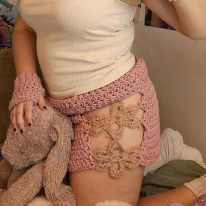 May include: A light pink crochet skirt with flower cutouts, matching fingerless gloves, and a white tank top. The skirt is adorned with a floral design. A small crochet fish is also visible.