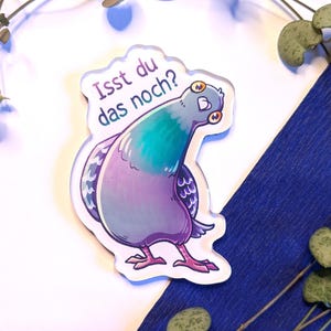 Op de afbeelding: Een sticker met een cartoon duif in teal, paars en wit. De duif heeft grote ogen en kijkt naar links. De Duitse tekst "Isst du das noch?" staat boven de duif. De sticker ligt op een witte en blauwe achtergrond.