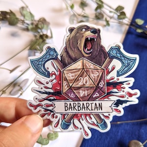 Könnte beinhalten: Aufkleber mit einem brüllenden braunen Bären, einem D20-Würfel und gekreuzten Äxten. Das Wort "BARBARIAN" steht auf einem Banner darunter. Rote Blutspritzer ergänzen das Design.