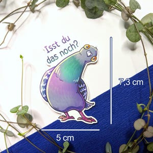 Puede incluir: Una pegatina troquelada con una paloma de dibujos animados con plumas iridiscentes moradas, verdes y azules. La paloma tiene grandes ojos amarillos y mira hacia atr&aacute;s. El texto "Isst du das noch?" est&aacute; impreso sobre el ave. Mide 7,3 cm por 5 cm.