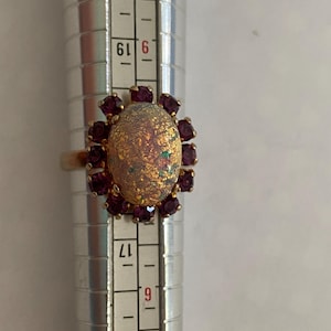 May include: A gold ring with an oval, iridescent opal-like stone, encircled by small, dark red gemstones. The ring is on a silver ring sizer. The design of the ring resembles a flower or sunburst.