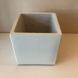 May include: A white square concrete planter. The planter is made of rough textured concrete and has a smooth white finish.