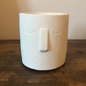May include: A white, cylindrical planter with a minimalist face design. The planter features a raised nose and closed eyes, giving it a serene appearance. The surface has a speckled texture, adding visual interest to the simple design.