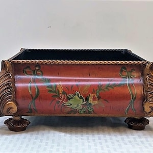 May include: A decorative planter with a red base and gold accents. The planter features a floral design with green leaves and pink and yellow flowers. The planter has a rope trim around the top and sits on four ornate feet.