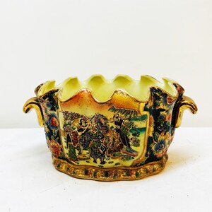 May include: A large, ornate ceramic planter with a scalloped rim and two gold handles. The planter is decorated with a colorful scene of people in traditional Japanese clothing, flowers, and trees. The planter has a gold trim and a glossy finish.
