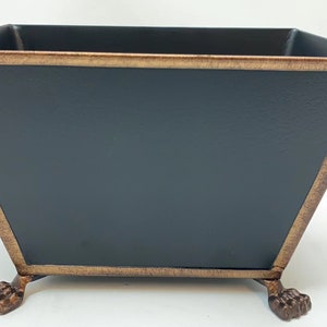 May include: A black metal planter with a gold-colored trim and decorative feet. The planter has a square shape with angled sides. The gold trim adds a vintage look to the planter.